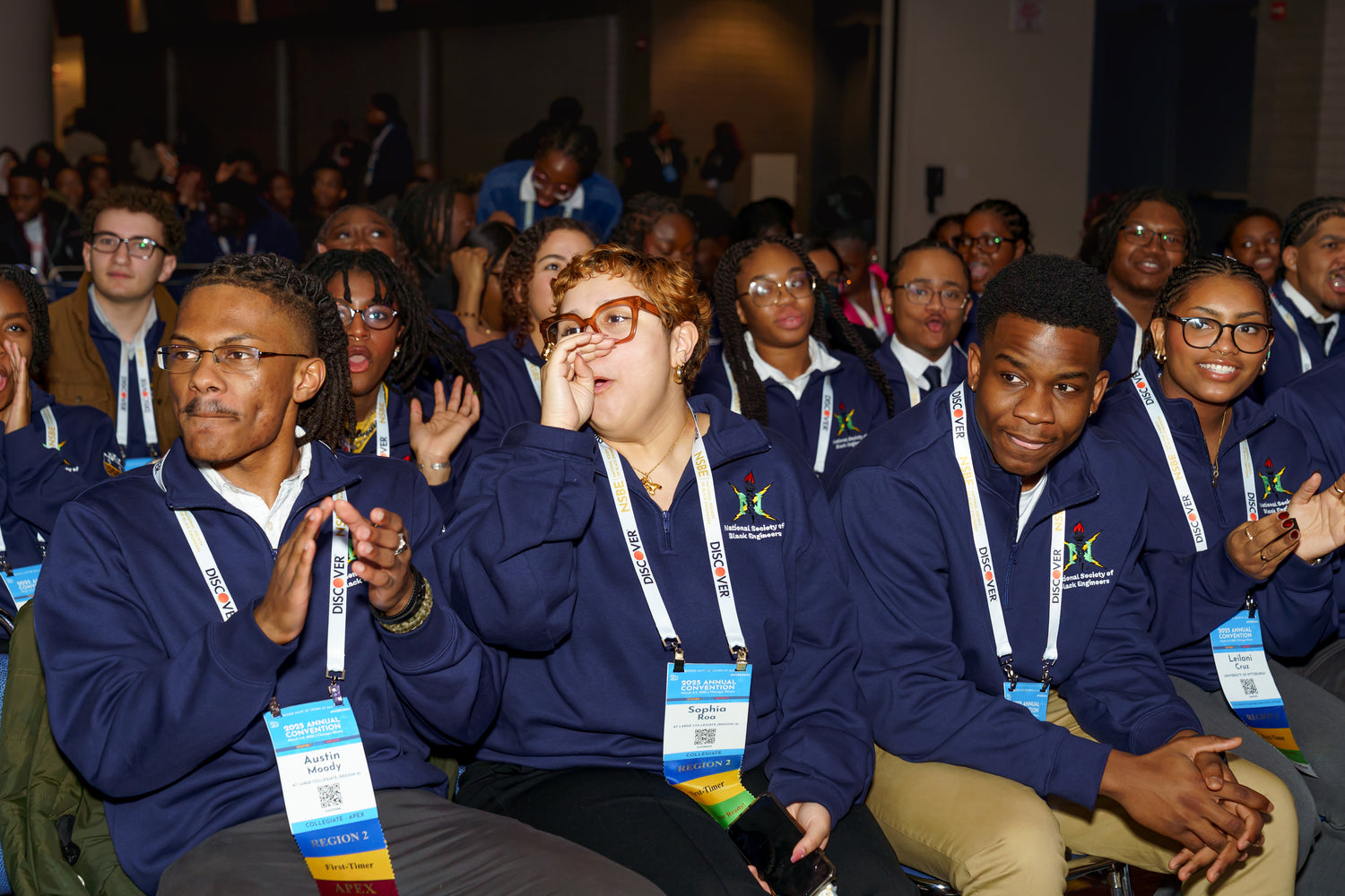 Group of people in blue jackets with lanyards, sitting and clapping in an indoor setting.